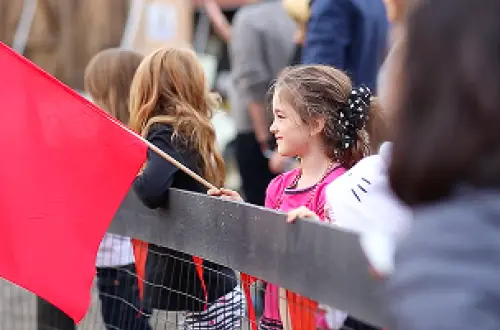 A child holds a red flag and watches an activity from behind a fence. 