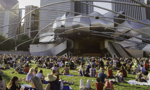 Hundreds of people sitting down on picnic rugs in front of Pritzker Pavillion 