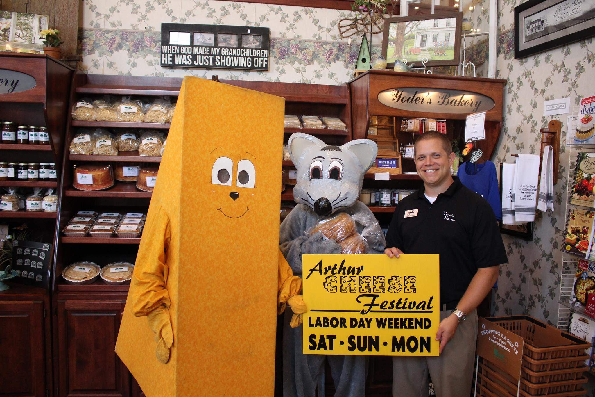 Mascots in a cheese costume and a mouse costume standing alongside a man holding an Arthur Cheese Festival sign