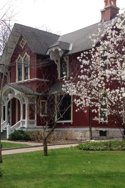A grand, two-storey, 19th century red wooden house, behind a lawn and blossom tree