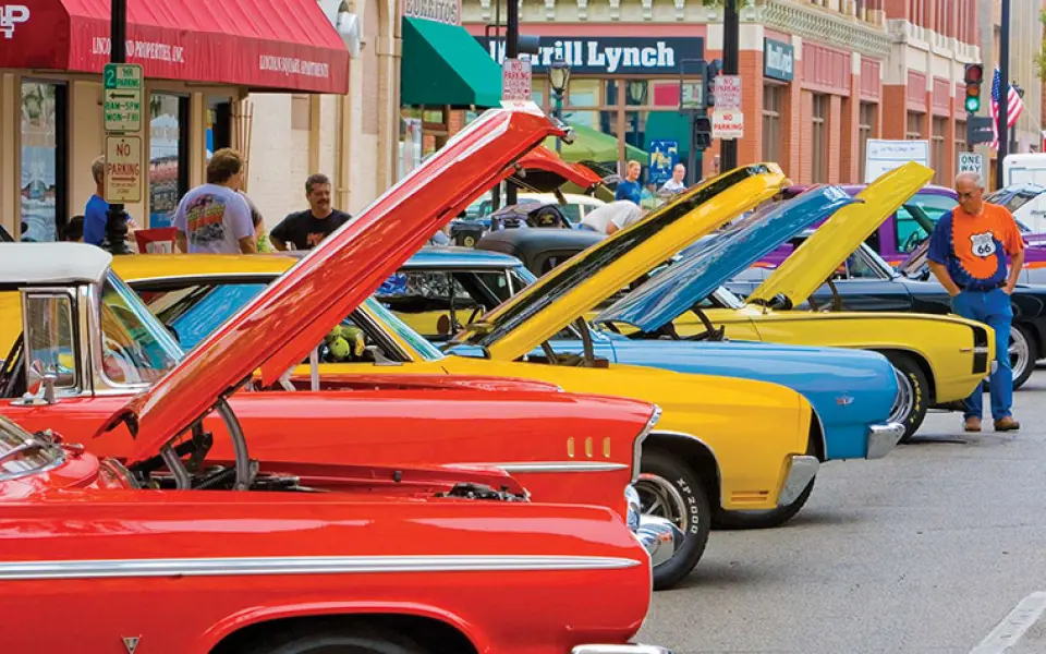 Colorful classic cars with their hoods raised line a downtown street during a Route 66 car show.