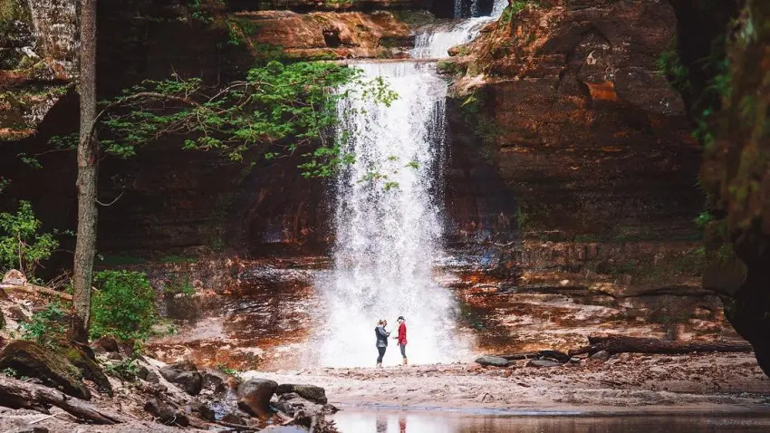 Two people standing in front of a waterfall, next to a serene pond.