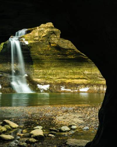Looking outwards from a cave, a waterfall cascades into a pool, next to snow-lined rock walls