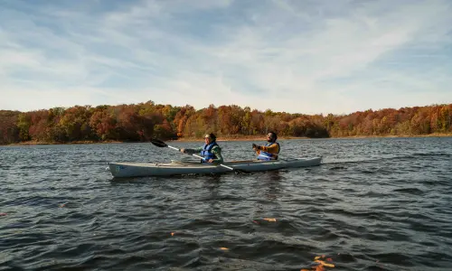 Two kayakers on a lake with fall foliage along the shore.