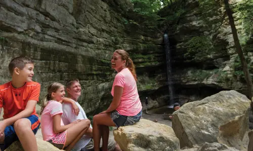 A family of four sitting down for a rest on rocks in front of a waterfall