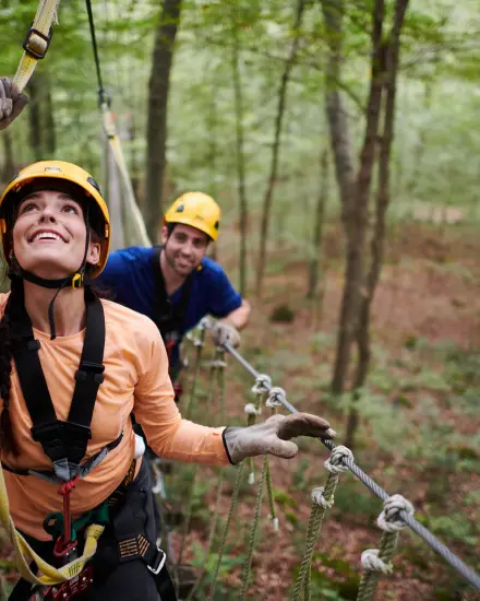 Two people wearing helmets and harnesses navigate an aerial ropes course in the woods.
