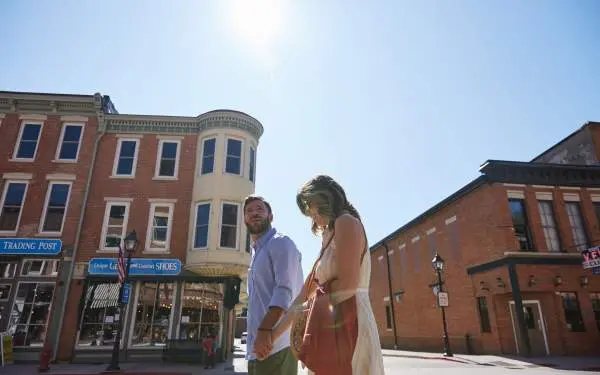 A low-angle shot of a young couple walking along a small town's main street, brick buildings and the sun behind them
