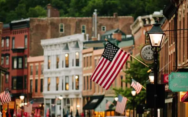 An American flag hanging from the front of a building on a small town main street full of brick buildings