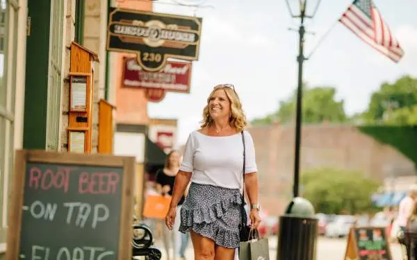 A woman walks down the sidewalk of the main street of a small town, looking in a store window. Old fashioned shop signs and an American flag hang above