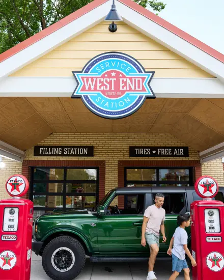 Father and son walk past a green SUV and vintage gas pumps at the West End Service Station on Route 66.