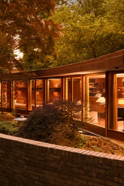 Modern curved glass house illuminated at dusk, surrounded by trees and a low brick wall.