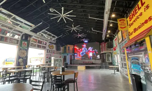 Interior of a spacious bar and live music venue with tables, vintage automotive signs and décor, and a large screen above the stage.