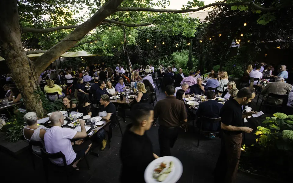 A crowded outdoor dining area at dusk, with tables amongst trees