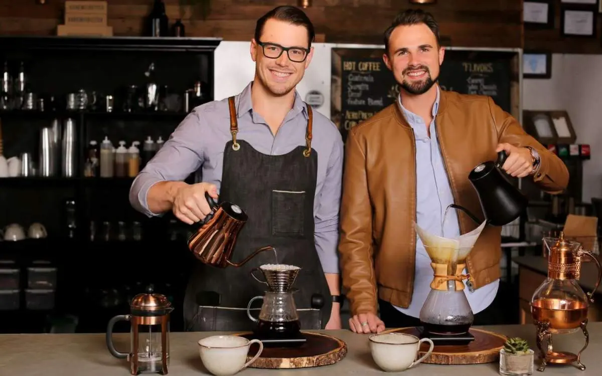 Two males looking into the camera while pouring filter coffee in a coffee shop