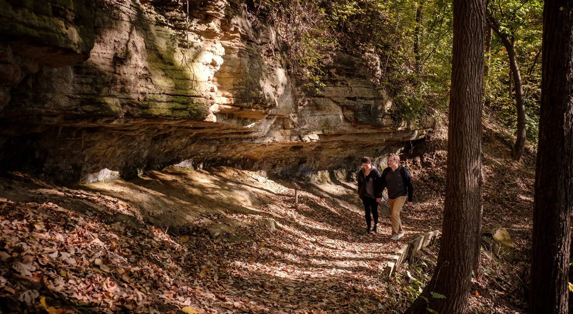 Two people walking in forest along a large rock