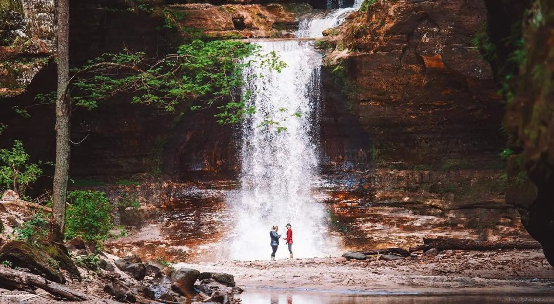 Two people standing in front of a waterfall, next to a serene pond.