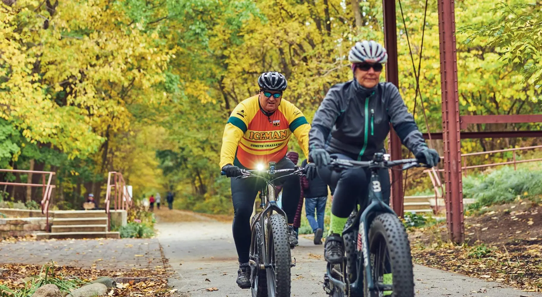 Two cyclists riding on a wooded trail beneath an old wooden bridge