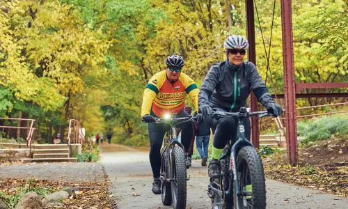 Two cyclists riding on a wooded trail beneath an old wooden bridge