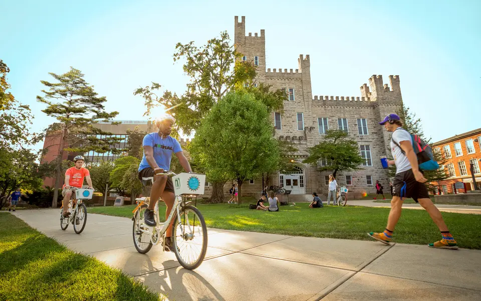 Two people bike through a university campus on a sunny day