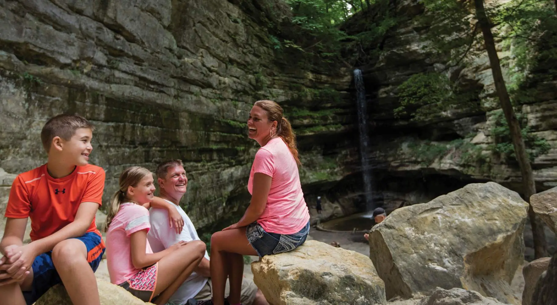 A family of four sitting down for a rest on rocks in front of a waterfall