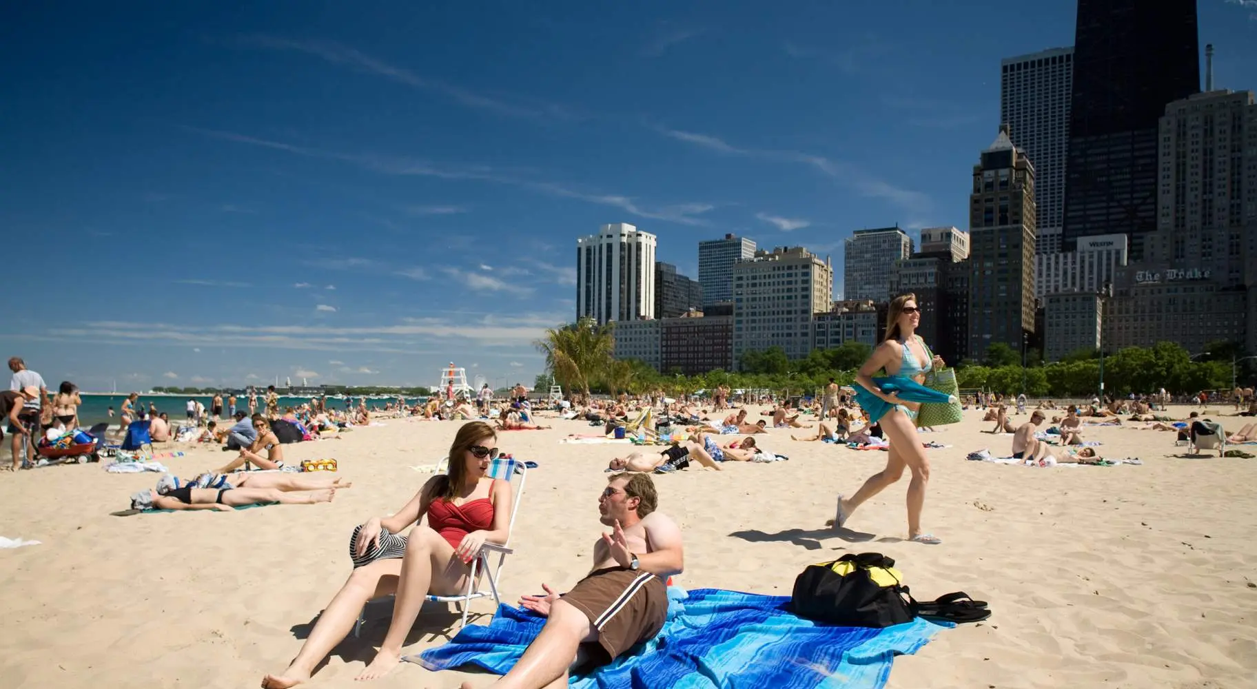 Two people lying on a sandy beach, with crowds in the background, along with the Chicago skyline