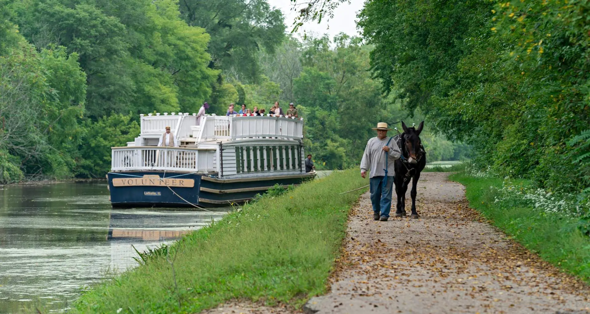 A boat going down the river and a man walking a Mule down a path