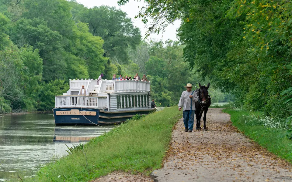 A boat going down the river and a man walking a Mule down a path